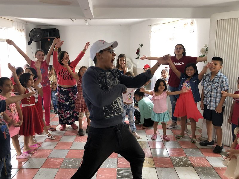 Many children watch a man with a white baseball cap perform in the center of a room.
