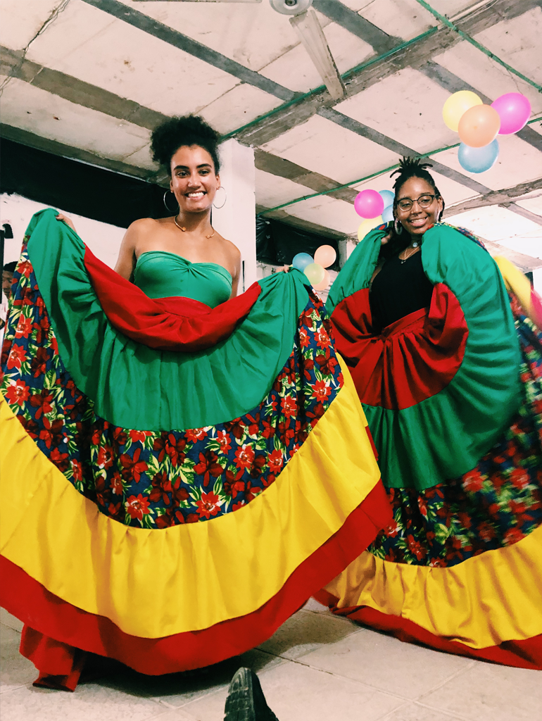 Two women wearing elaborate dresses that are red, gold, and green with floral pattern accents.