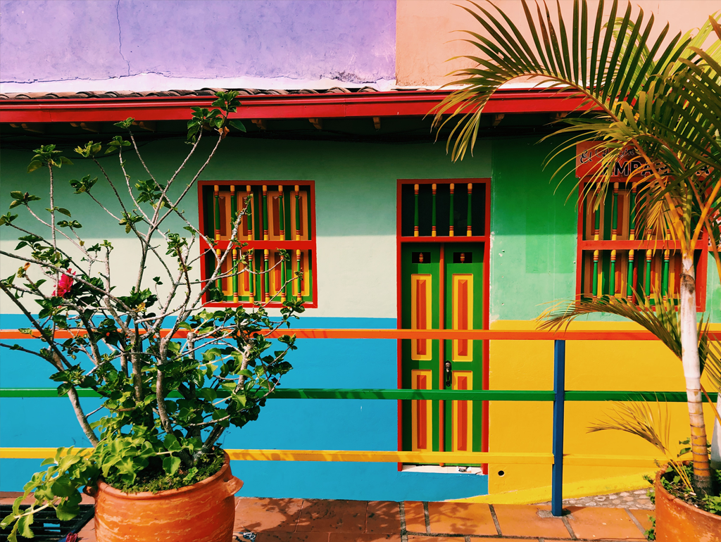 A fence painted many different colors. In the background is a house painted blue, green, yellow, and white.