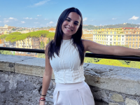 Maria Martinez posing in front of a stone wall with colorful buildings, green trees, and a blue sky in the background