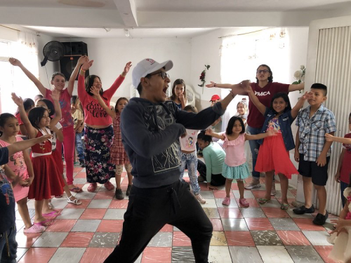 Many children watch a man with a white baseball cap perform in the center of a room.