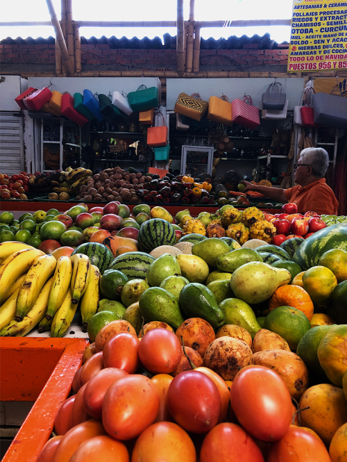 A gray-haired man with glasses tends a large fruit stand containing many different fruits.