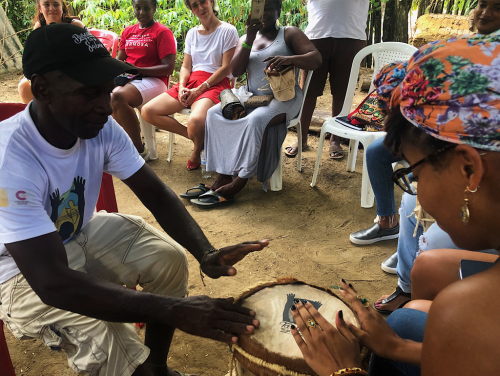 A man and woman play a drum with their hands while sitting with others in a circle of chairs.