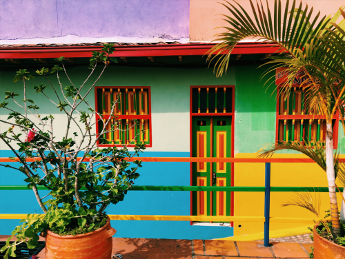 A fence painted many different colors. In the background is a house painted blue, green, yellow, and white.