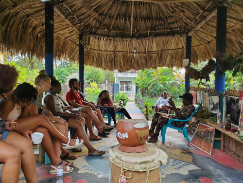 Students sit in chairs while listening to another sitting man speak underneath a pavilion with a thatched roof.